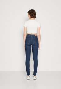Jeans de ganga skinny em azul escuro, com uma cintura alta, bolsos traseiros e uma etiqueta da marca castanha na cintura. Combinados com um top curto branco.
