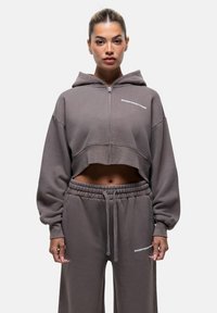 Felpa grigia cropped con zip, maniche over-size e testo ricamato. Abbinata a pantaloni della tuta grigi coordinati con una vita elasticizzata.