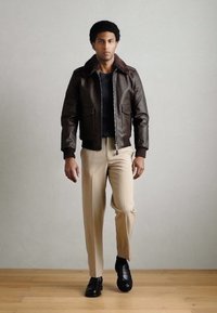 Barna bőr bomber dzseki műszőrme gallérral, krémszínű nadrággal és fekete cipővel párosítva; cipzáras záródással és elülső zsebekkel.