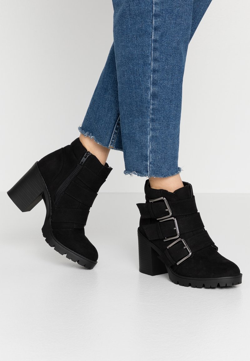Bottes à talons noires à la cheville avec trois boucles en argent portées avec un jean bleu raccourci sur un fond clair.