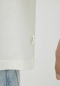 T-shirt blanc en coton avec une petite étiquette "BZB" en or, présentant une coupe droite et un détail de couture à l'ourlet, associé à un denim bleu.