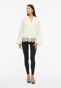 Lys beige skjorte med knapper og vide ermer, med blondedetalj nederst, brukt over svarte leggings og sko med spiss tå.