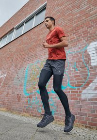 Corridore in maglietta atletica rossa, leggings neri e scarpe nere, che corre su un marciapiede accanto a un muro di mattoni texturizzati con graffiti.