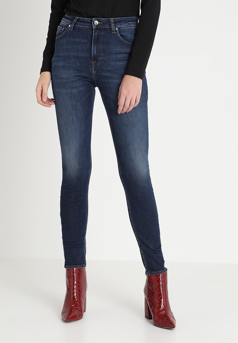 Jeans oscuros ajustados de talle alto y diseño clásico de cinco bolsillos, combinados con botas de tobillo texturizadas rojas.