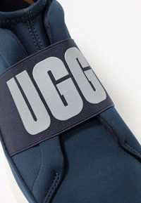Granatowe sneakersy wsuwane, wykonane z elastycznego materiału, z szerokim czarnym paskiem z jasnoszarym logo "UGG" oraz gładkim, zaokrąglonym kształtem.