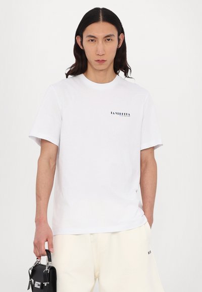 T-shirt en coton blanc à manches courtes, avec un petit logo noir sur le devant. Le modèle tient un sac noir avec une poignée.