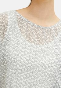 Gros plan sur une personne portant une chemise blanche avec un motif répétitif de vagues en demi-cercle noires et un col arrondi.