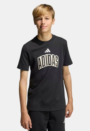 Ragazzo adolescente con capelli biondi corti che indossa una t-shirt nera Adidas e pantaloni neri, in piedi davanti a uno sfondo chiaro e uniforme.