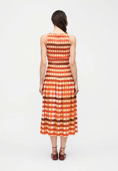 Γυναίκα που στέκεται γυρισμένη προς τα πίσω, φορώντας αμάνικο καρό φόρεμα midi σε πορτοκαλί, ροζ και καφέ χρώμα, με μπορντό δερμάτινα παπούτσια με κορδόνια, σε λευκό φόντο.