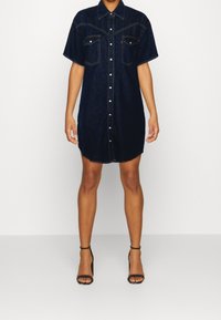 Mörk denim oversized skjortklänning med korta ärmar, knappstängning, två bröstfickor och rundad nederkant. Bärs med svarta sandaler med remmar.