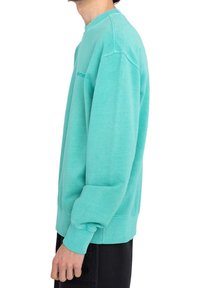 Homme portant un sweat-shirt turquoise à col ras du cou et un pantalon noir, debout de profil avec le bras gauche détendu, visible du cou jusqu'aux hanches.