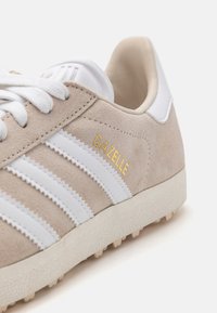 Sneakers en suède beige clair avec des accents en cuir blanc, dotées de trois rayures blanches et d'un logo "GAZELLE" doré. Semelle en caoutchouc texturée avec des clous.