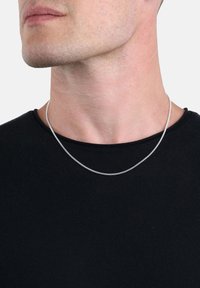 Primo piano del volto inferiore e del collo di una persona che indossa una sottile collana d'argento e una maglietta nera a collo rotondo, su uno sfondo bianco.