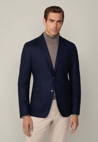 Blazer di colore blu navy con due bottoni, rever a punta, tasche frontali e texture liscia. Indossato sopra un maglione a collo alto marrone chiaro.