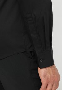 Camicia nera a maniche lunghe, con polsini abbottonati, texture liscia, vestibilità su misura e dettagli delle cuciture lungo il braccio e l'orlo.