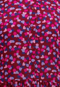 Czerwona sukienka z tkaniny, zdobiona sercowym wzorem w jasnoróżowym, fioletowym i niebieskim kolorze, z zapięciem na guziki i falbaną u dołu.