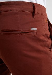 Pantaloni color ruggine con una texture liscia, tasche frontali e posteriori, e un'etichetta metallica logo attaccata alla tasca posteriore.