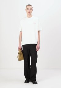 T-shirt blanche à manches courtes avec texte rouge sur le devant, associée à un pantalon cargo noir ample. Le modèle tient un objet beige plié dans une main.