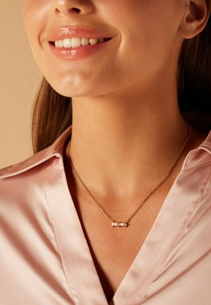 Collier en or avec trois petites pierres rondes. Porté sur une chemise en satin rose pâle, mettant en valeur le design délicat du collier.