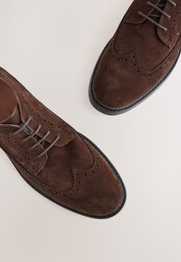 Chaussures brogues en daim marron avec une finition texturée, présentant un design classique à bout ailé et des semelles en caoutchouc noir. Fermeture à lacets avec des perforations décoratives.