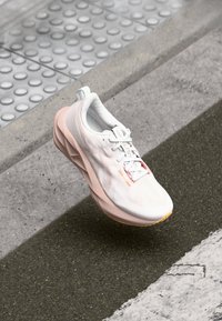 Zapatilla de running ligera en blanco y rosa suave, con un upper texturizado, suela acolchada y detalle de logo acentuado en el costado.