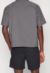 Šedá košile s krátkým rukávem, rovný lem a detaily na zadním díle, spárovaná s černými šortkami. Tkanina má hladkou texturu, uvolněný design.