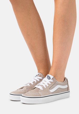 Paar beige Vans-Sneaker mit weißen Schnürsenkeln und weißem Seitenstreifen, getragen barfuß vor neutralem Hintergrund.