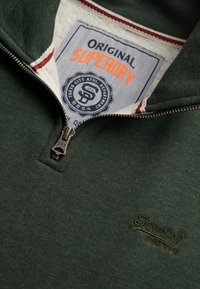 Donkergroene pullover met een kwart ritssluiting, vervaardigd van een textuurstof, met contrasterende kraagstrepen en een logo patch op de borst.