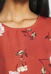 Top floreale rosso con scollatura rotonda, tessuto testurizzato, con motivi floreali rosa e marroni su uno sfondo liscio.
