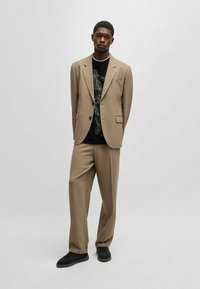 Beige suit with a single button closure and two front pockets, worn over a black graphic t-shirt. Black sneakers complete the outfit.