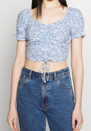 Blaues Blumen-Crop-Top mit Puffärmeln, gerafften Details und verstellbarem Knoten am Saum, kombiniert mit hochtaillierten Jeans.