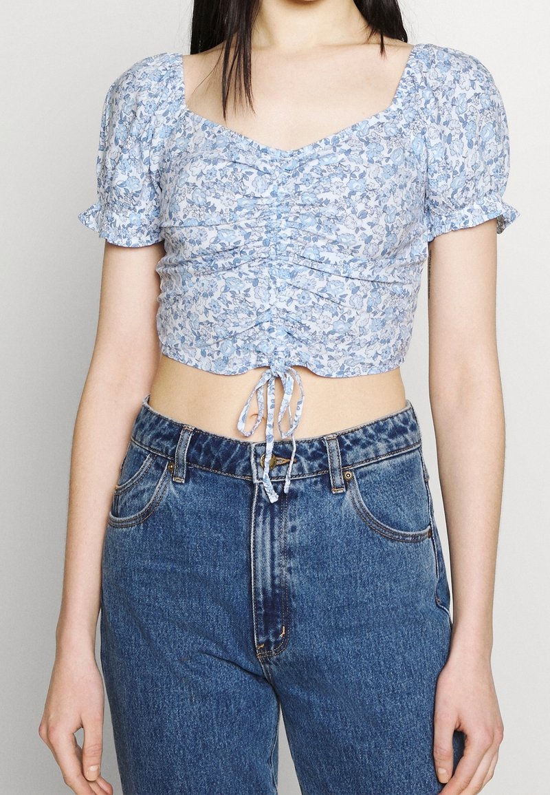 Top corto floral azul con mangas abullonadas, detalles fruncidos y un lazo ajustable en el dobladillo, combinado con vaqueros de cintura alta.