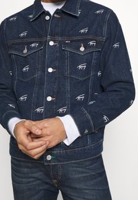 Jeansjacka i mörkblått med ett broderat logomönster. Har knappstängning och två bröstfickor med silverdetaljer.