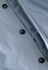 Țesătură gri deschis, cu o textură netedă, decorată cu nasturi închis la culoare și cusături contrastante. Vedere de aproape care evidențiază amplasarea detaliată a nasturilor.
