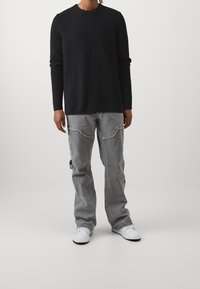 Zwart getextureerd brei-sweater met lange mouwen, gecombineerd met lichtgrijze cargobroek met zakken en rafelige randen. Witte sneakers maken de look compleet.