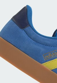 Sneaker in suede blu con strisce gialle, tallone blu scuro e suola in gomma testurizzata. Ingrandimento del materiale superiore e degli elementi di design.