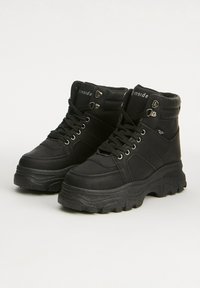 Par de botas altas de cuero negro con suelas gruesas, ojales de metal, cuellos acolchados y cordones negros sobre un fondo claro y liso.