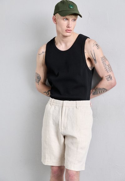 Schwarzes geripptes Tanktop, beige Leinen-Shorts mit Falten und eine grüne Kappe mit einem bestickten Patch. Glatte Textur, lässiges Design.