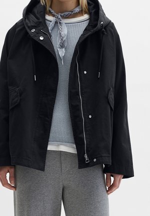 Person trägt eine schwarze Kapuzenjacke über einem blauen Strickpullover, graue Hose und ein hellblau gemustertes Halstuch, das zu einer Schleife gebunden ist.