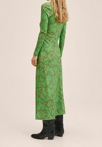 Langärmliges grünes Kleid mit einem Kettenmuster in Goldtönen, glatter Textur und knöchellangem Saum, kombiniert mit schwarzen Absatzstiefeln.