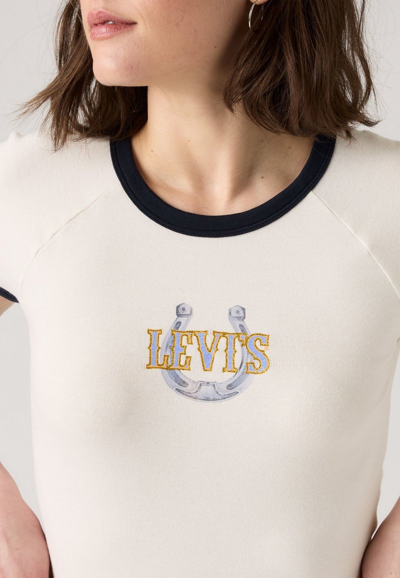 Kurzarmliges cremefarbenes Oberteil mit einem navyblauen Rippsaum, das den gestickten Schriftzug "LEVI'S" und ein grafisches Hufeisen-Design in Grau und Gelb zeigt.