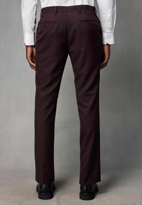 Burgundske krojene hlače s glatkom teksturom, slim fit krojem i dva stražnja džepa zatvorena crnim dugmadi, uparene s crnim cipelama.