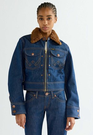 Junge Frau mit geflochtenem Haar trägt dunkelblaue Jeansjacke mit braunem Lammfellkragen und passende Jeans vor weißem Hintergrund.