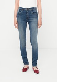Jeans de mezclilla azul ajustados con zonas desgastadas, diseño de cinco bolsillos y cierre de botón. Llevados con zapatos planos rojos.