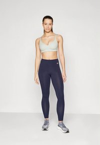 Žena stojící v lehce šedém sportovním topu, tmavě modrých legínách s vysokým pasem a šedých sportovních botách na bílém pozadí.