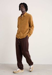 Hemd met lange mouwen in mosterdkleurige corduroy, met een borstzakje, gecombineerd met loszittende bruine broek en veelkleurige sneakers.