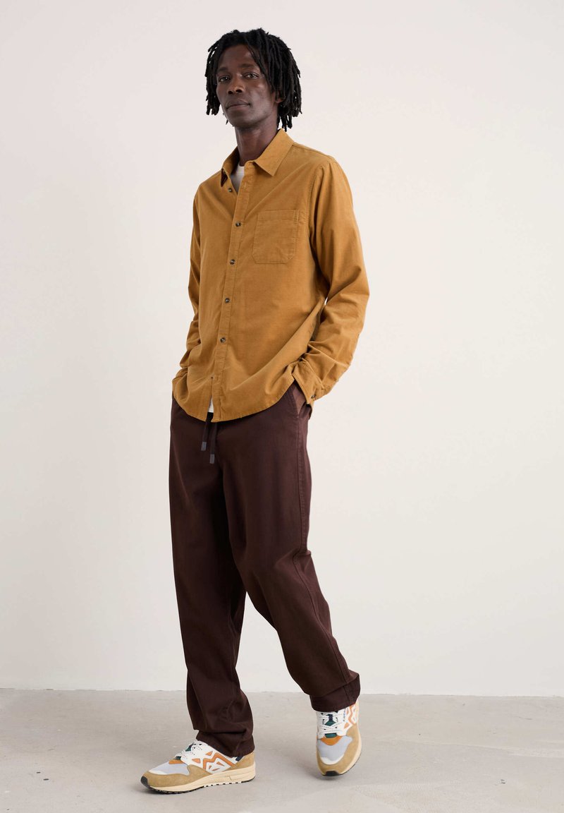 Hemd met lange mouwen in mosterdkleurige corduroy, met een borstzakje, gecombineerd met loszittende bruine broek en veelkleurige sneakers.