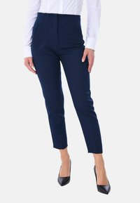 Pantaloni tailored navy con vita alta, caratterizzati da pieghe, tasche laterali e gamba slim-fit, abbinati a décolleté nere con punta affusolata.