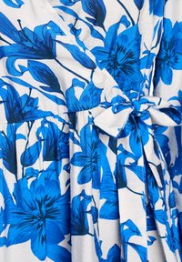 Sinine lilleline mähkmekleit, kus on suured sinised liiliad valgel taustal, millel on vöökoht ja kokkuvõetud seeliku detail.