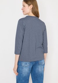 Femme aux cheveux châtain clair portant un haut à manches longues à rayures verticales bleu marine et blanc et un jean bleu, de profil avec un léger sourire.